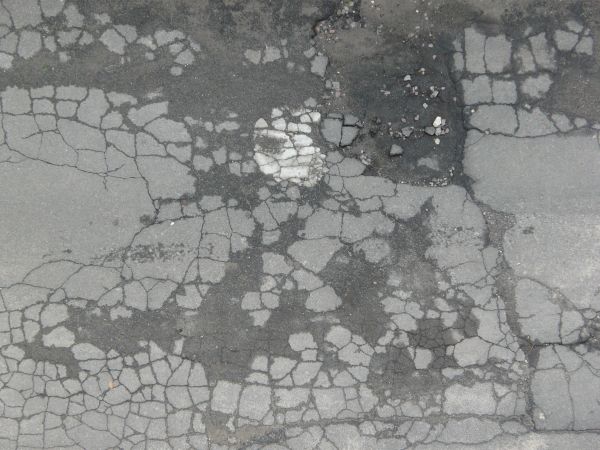 Cracked asphalt texture with many dark, damp patches, and a pothole filled with dark mud and small rocks.